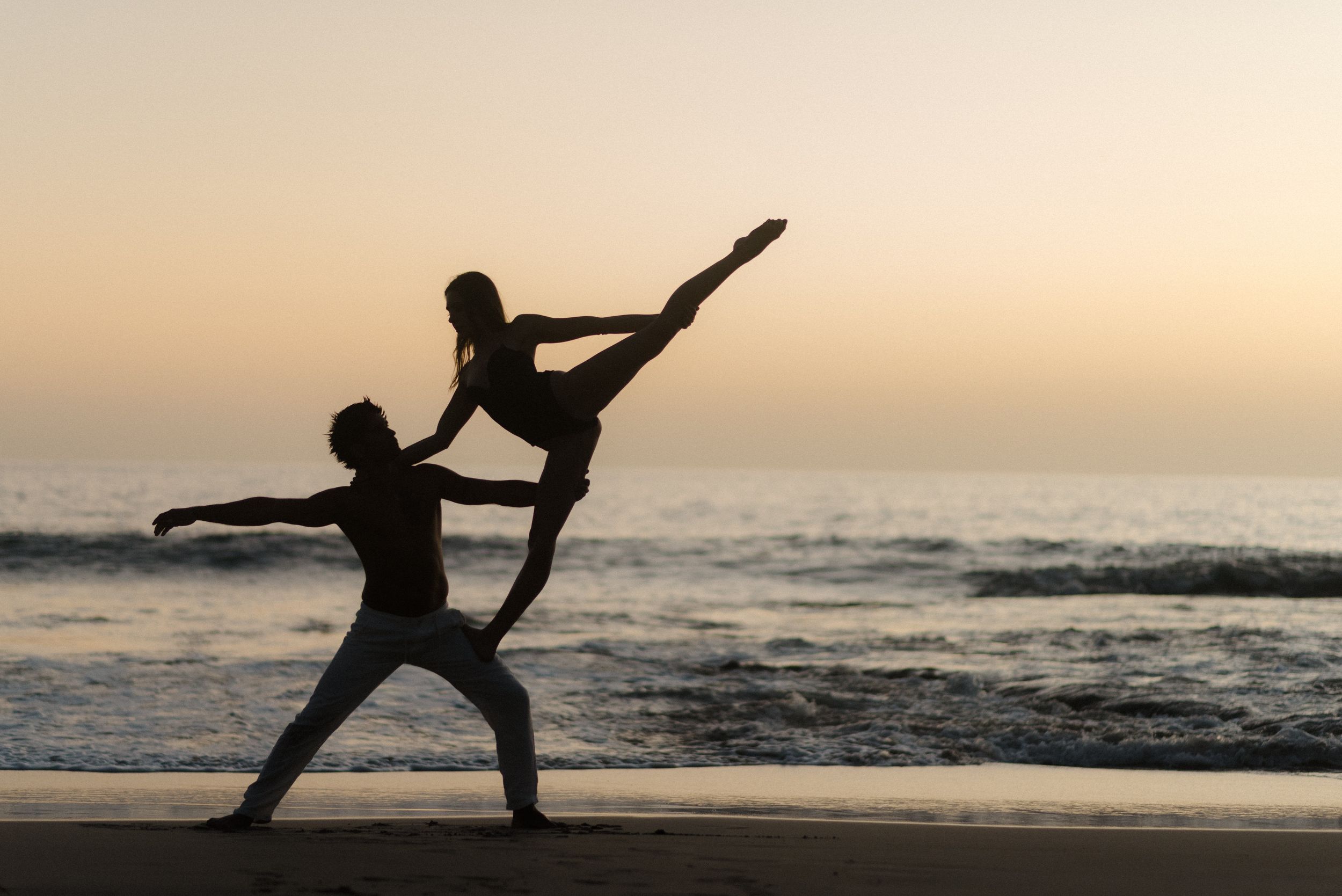 Silhouetted dancers perform an elegant ballet pose against a sunset beach backdrop with ocean waves.