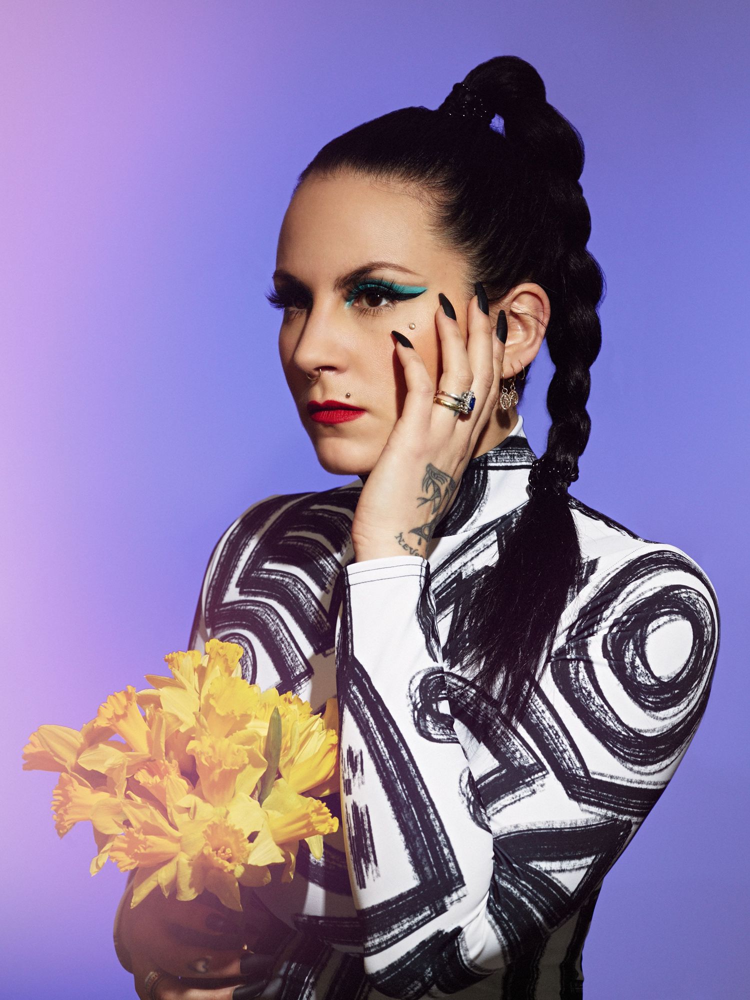 Person with dark braided hair, bold makeup, and tattoos wearing a graphic top while holding yellow flowers.