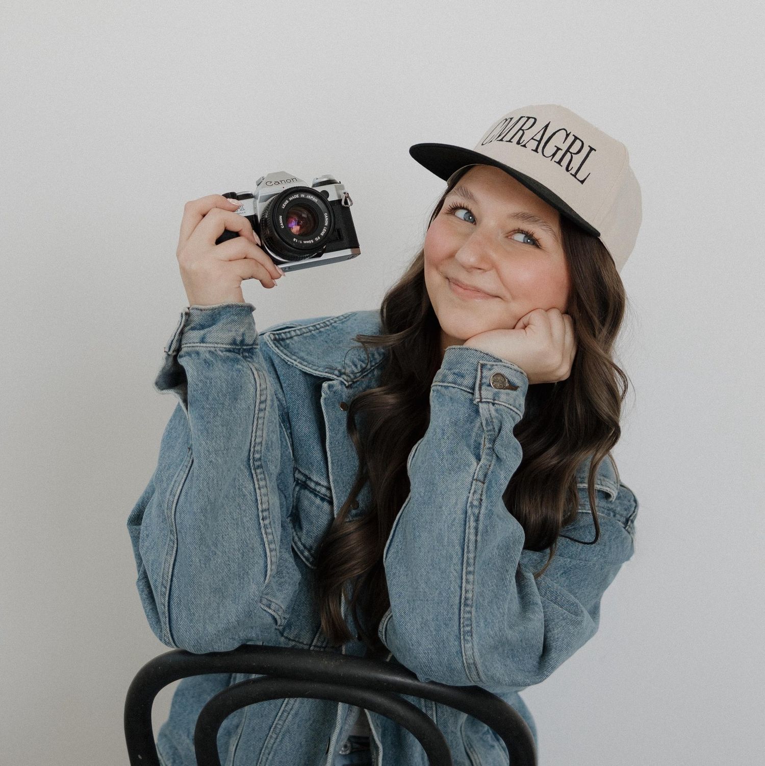 Young woman in denim jacket and OURAGRL cap holds a vintage film camera, smiling against a white background.