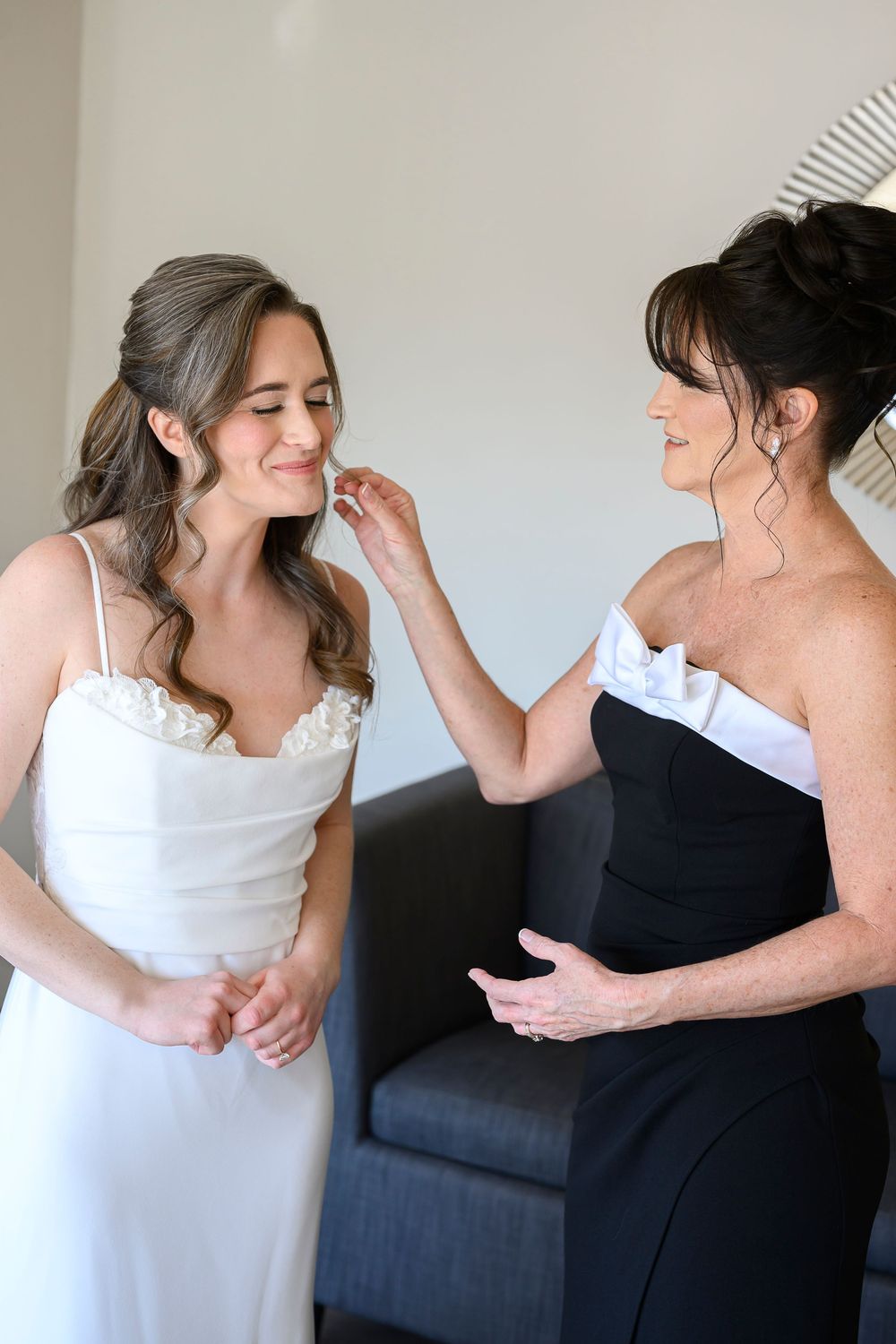 Bride in white gown smiles as older woman in black dress gently touches her face during wedding preparations.