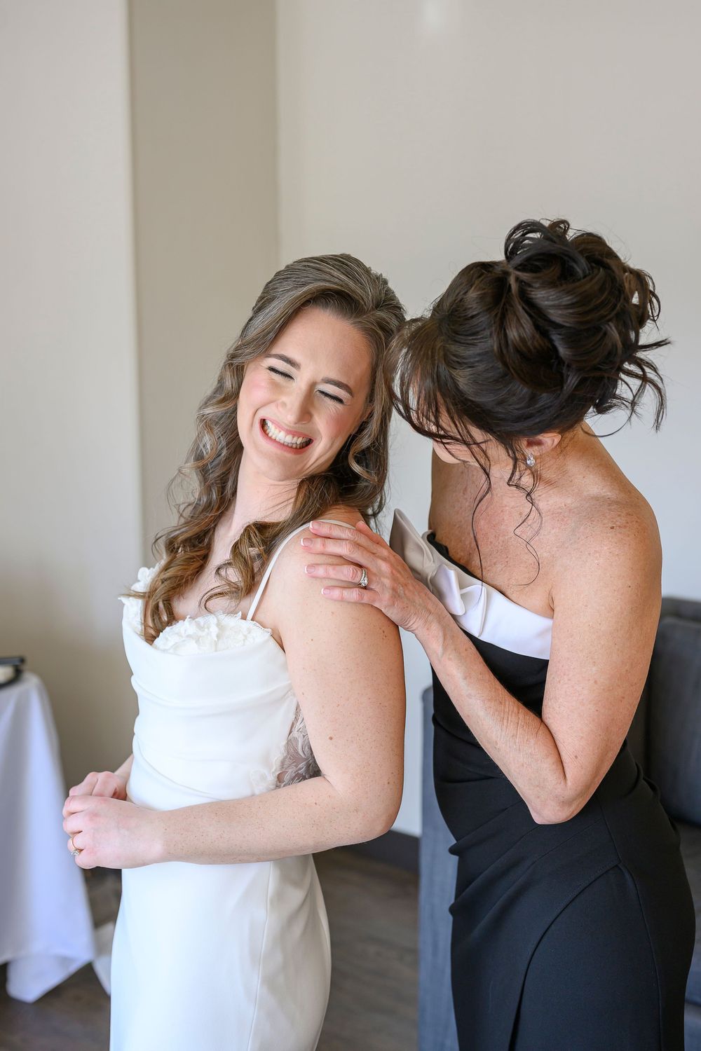Bride in white dress laughs joyfully as bridesmaid in black dress helps her get ready before the wedding ceremony.