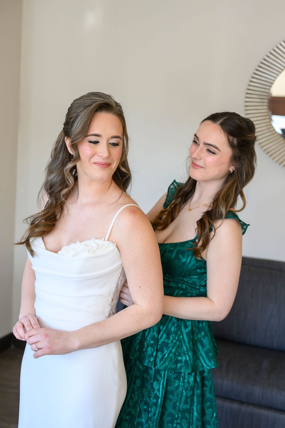 A bridesmaid in a green dress helps a bride in a white gown button her dress before the wedding ceremony.