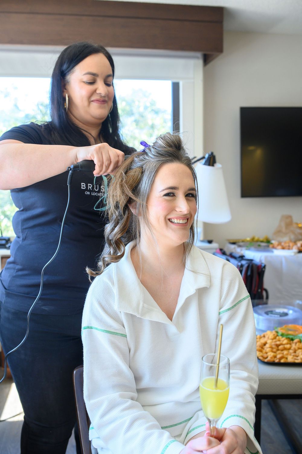 Bride smiling in white robe holds mimosa while hairstylist styles her hair during wedding morning preparations.