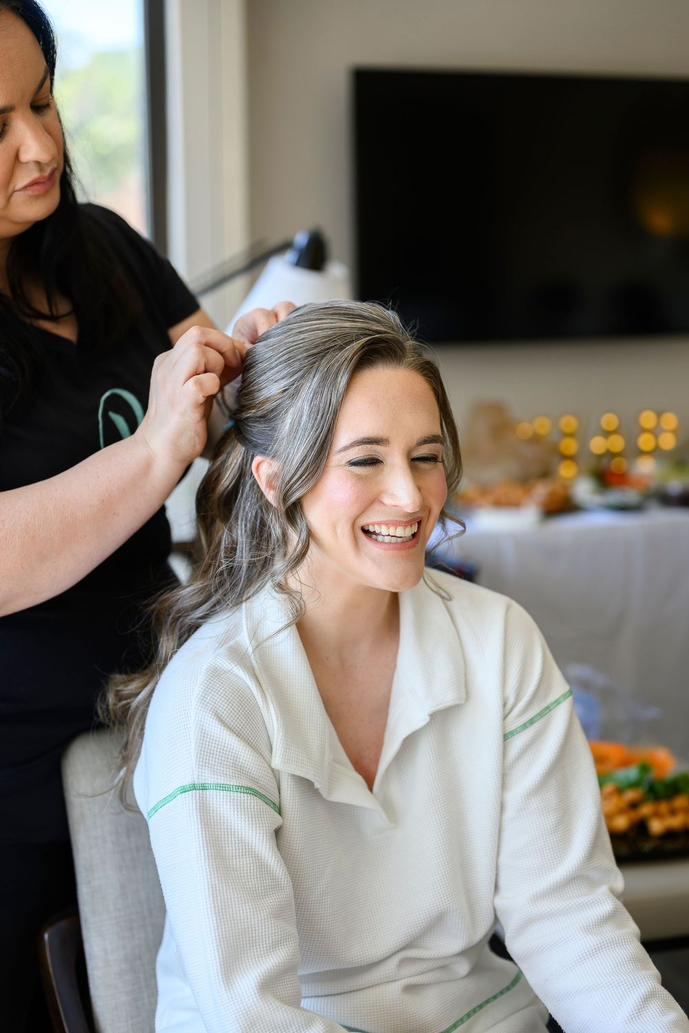 Bride smiling joyfully while hairstylist styles her gray-streaked hair during wedding preparations indoors.