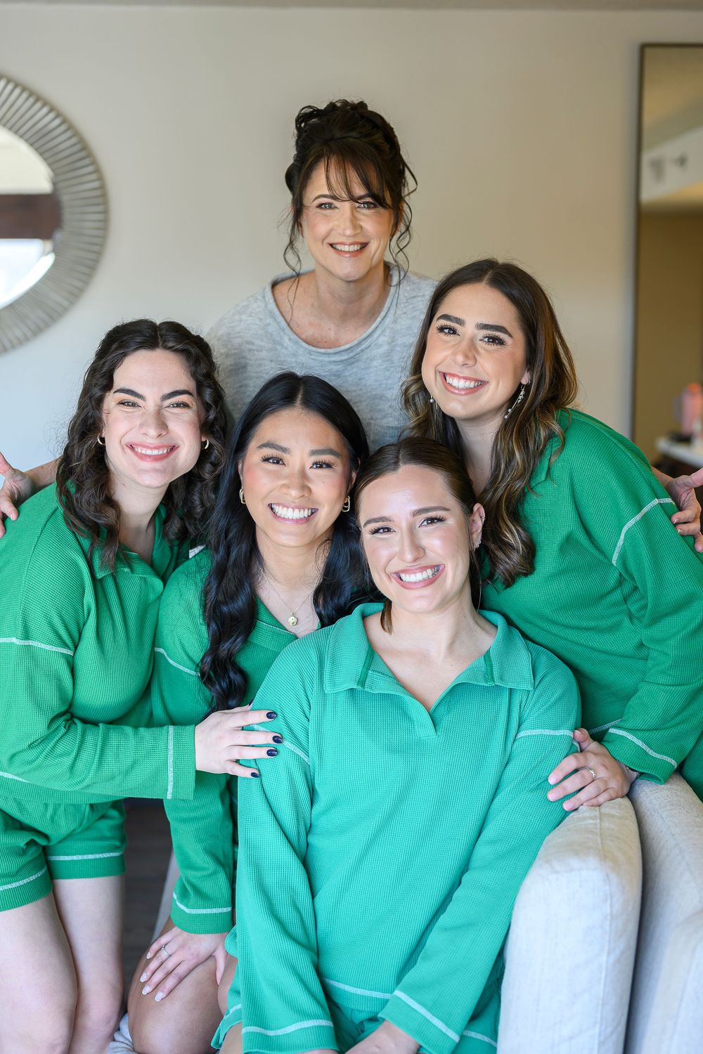 Five women in matching green pajamas smile together in a bright room during a bridal party getting-ready session.