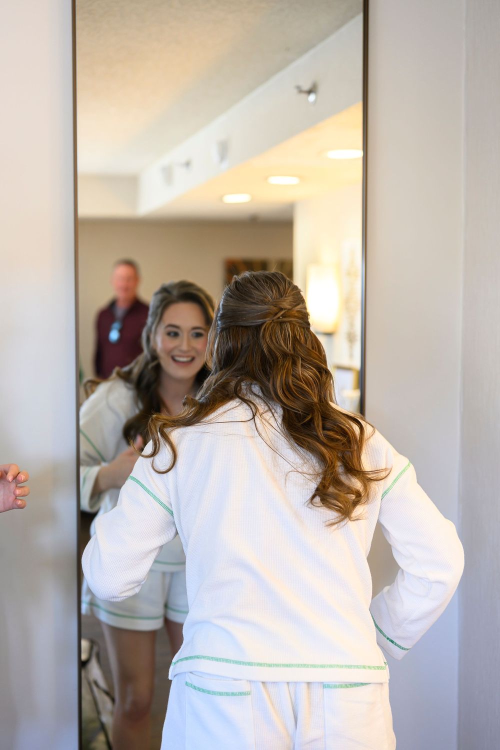 Bride in white pajamas with green trim admires her half-up hairstyle in a full-length mirror before her wedding.
