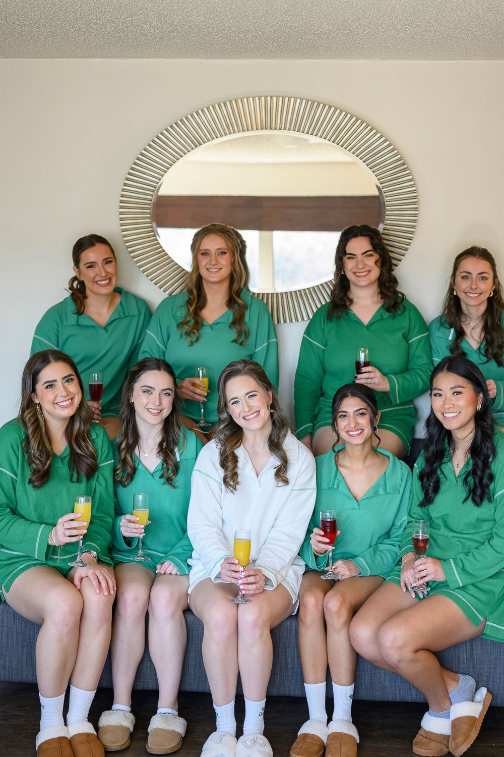 Bride in white robe with bridesmaids in green pajamas holding champagne glasses, posing together in a hotel room.