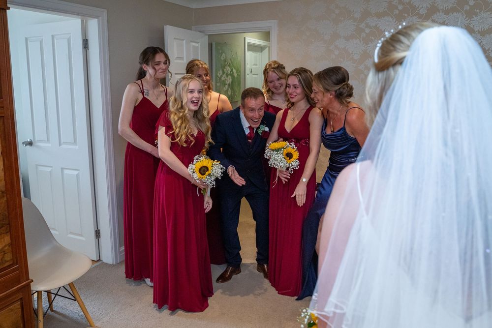 Bridesmaids in red dresses and groomsman react joyfully as bride reveals her wedding dress.
