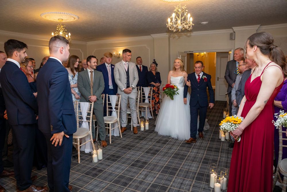 Bride walks down the aisle in tartan-floored ceremony room toward groom and waiting wedding guests.