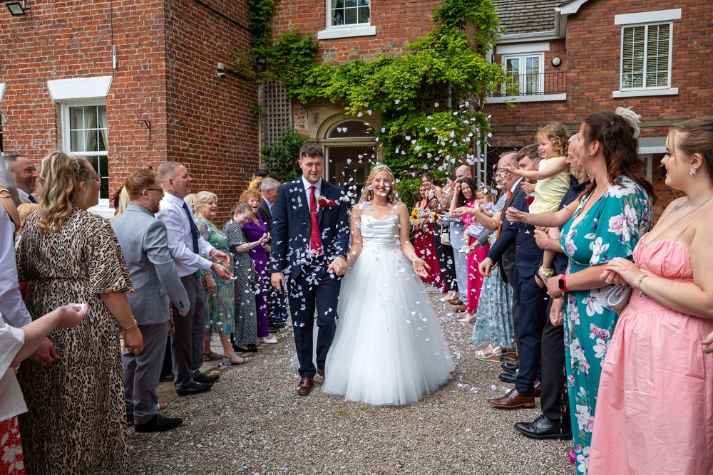 Newlyweds walk through confetti shower thrown by smiling guests outside a brick wedding venue.
