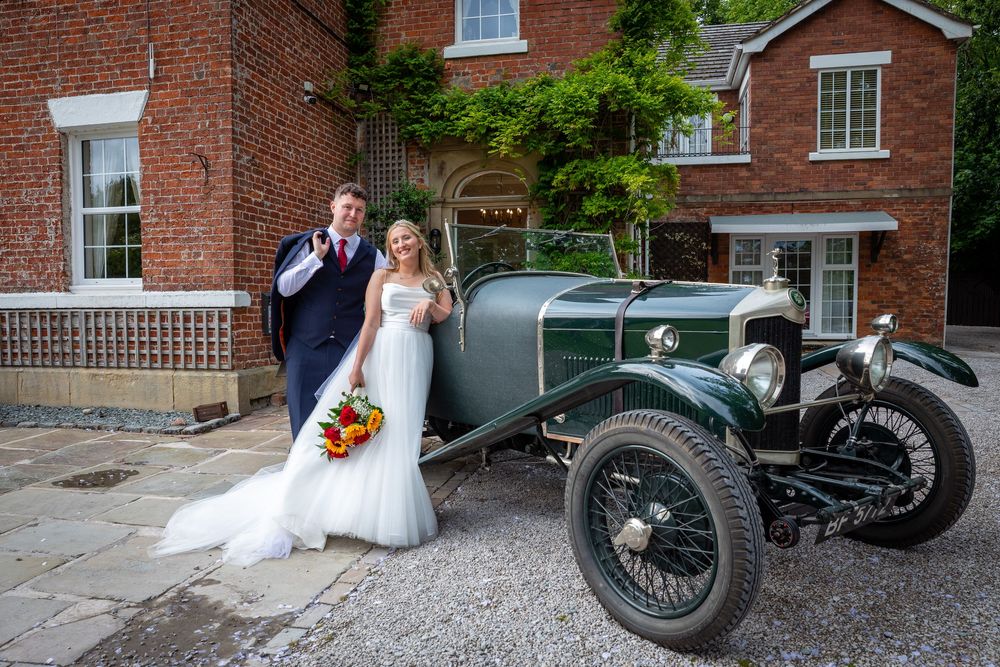 Smiling bride and groom pose beside a vintage green classic car outside a brick wedding venue.