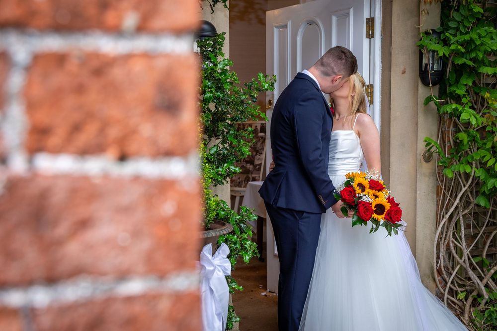 Groom kisses bride holding vibrant sunflower bouquet in doorway decorated with white ribbon and greenery.