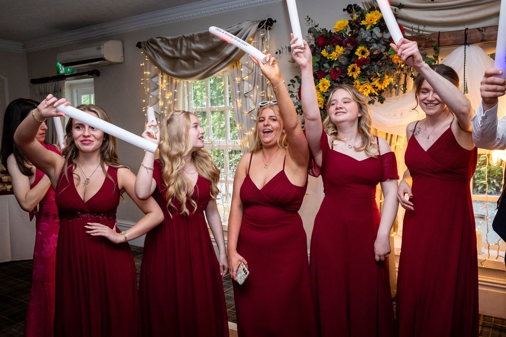 Bridesmaids in red dresses raise white scrolls and cheer during a wedding reception celebration.