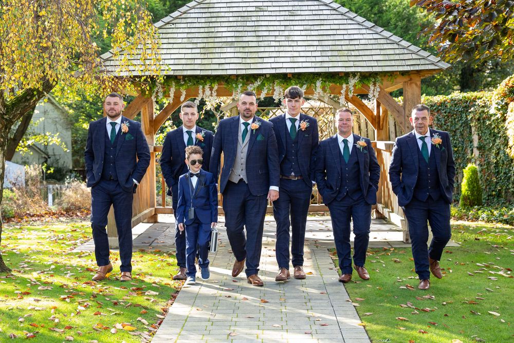 Groomsmen in navy suits walk together along garden path with wooden gazebo in autumn setting.