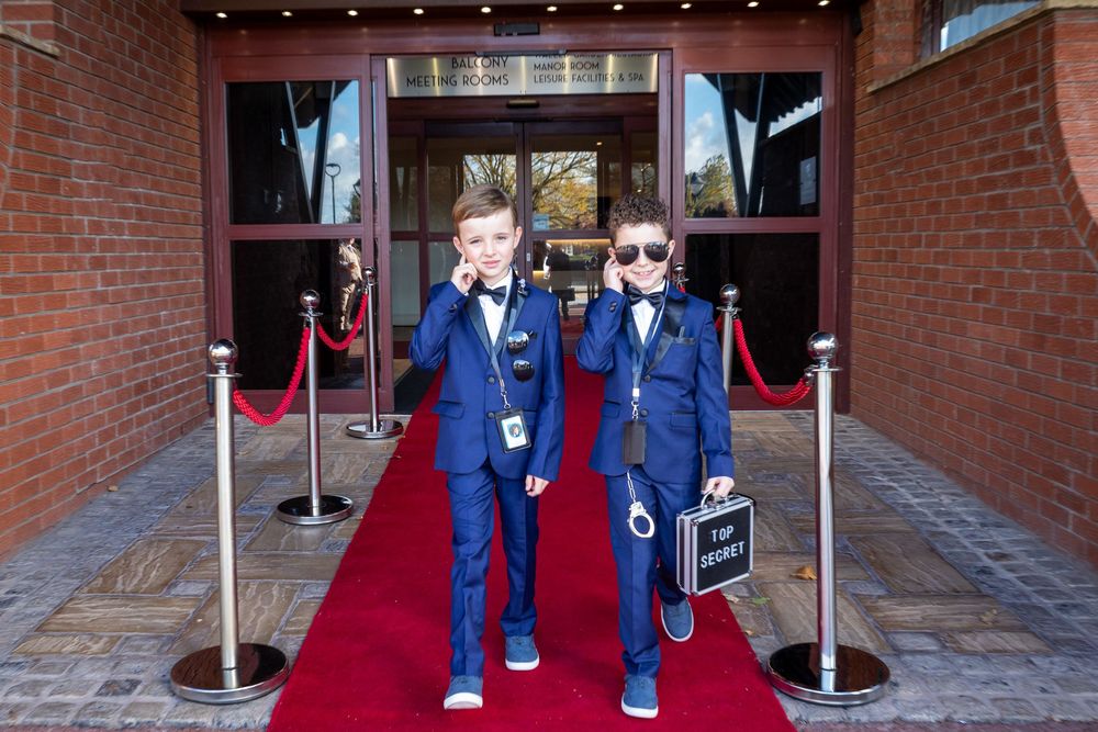 Two young boys in navy suits pose on red carpet entrance holding walkie-talkies and briefcase.