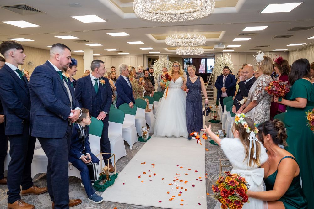 Bride walks down flower-petal aisle in elegant grey gown past smiling guests in decorated ceremony room.