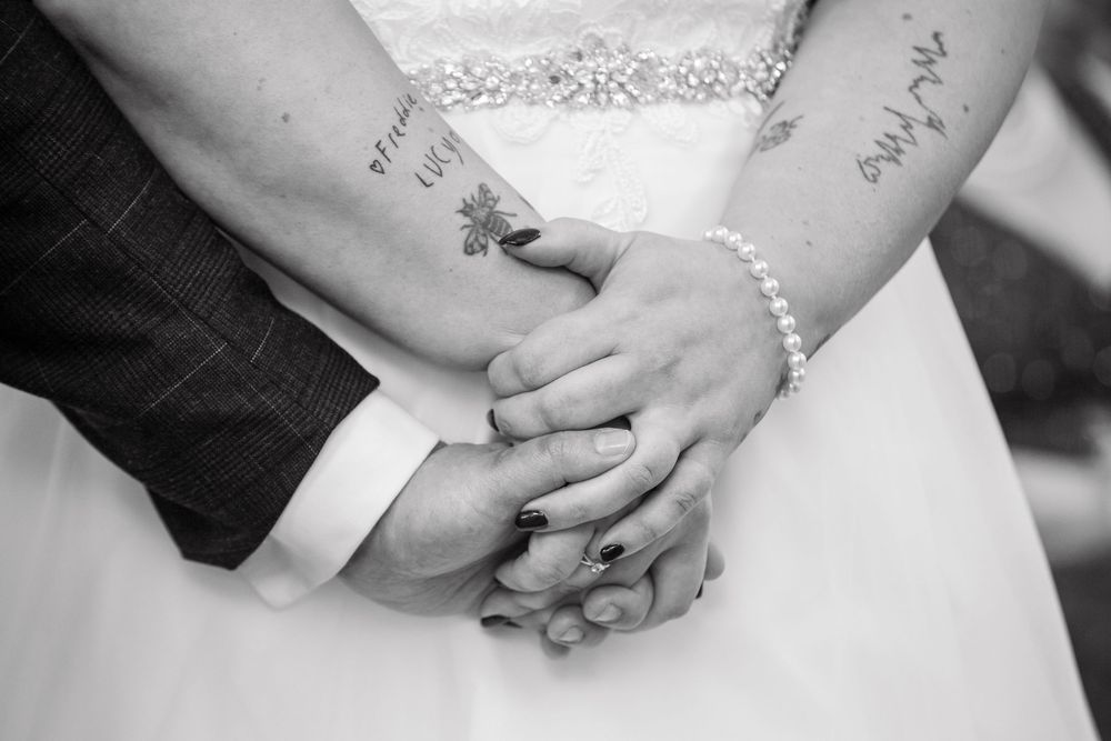 Black and white close-up of bride and groom holding hands showing tattoos and pearl bracelet.