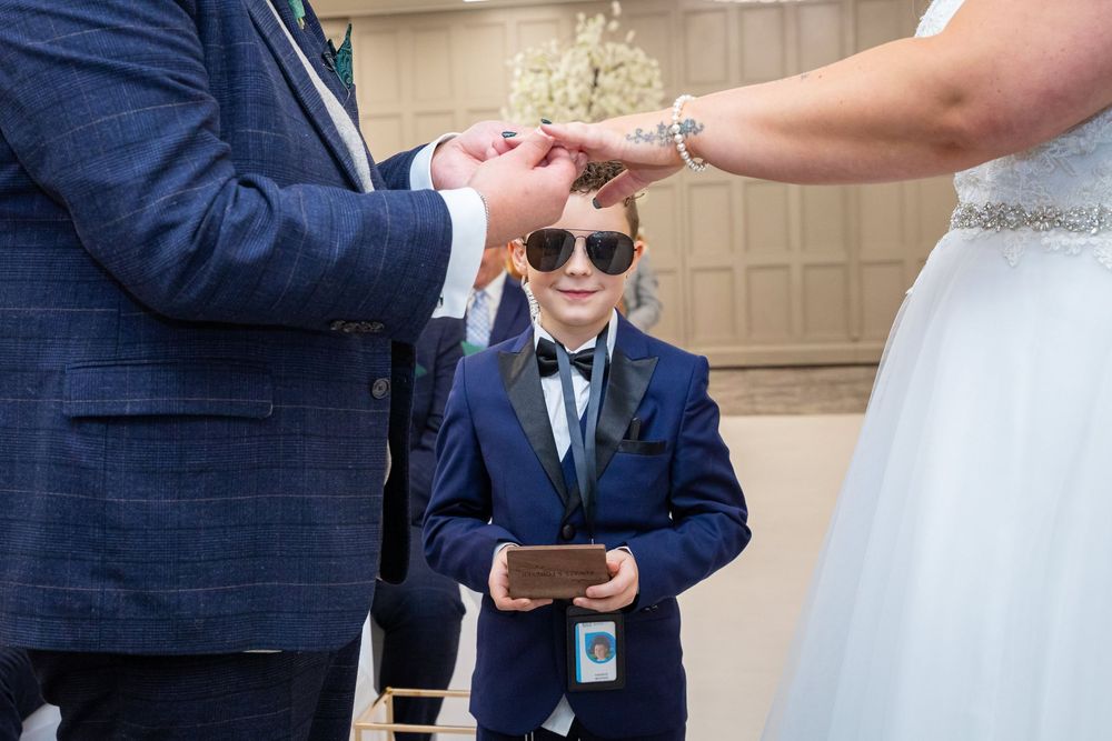 Young ring bearer in sunglasses and navy suit watches couple exchange rings during wedding ceremony.