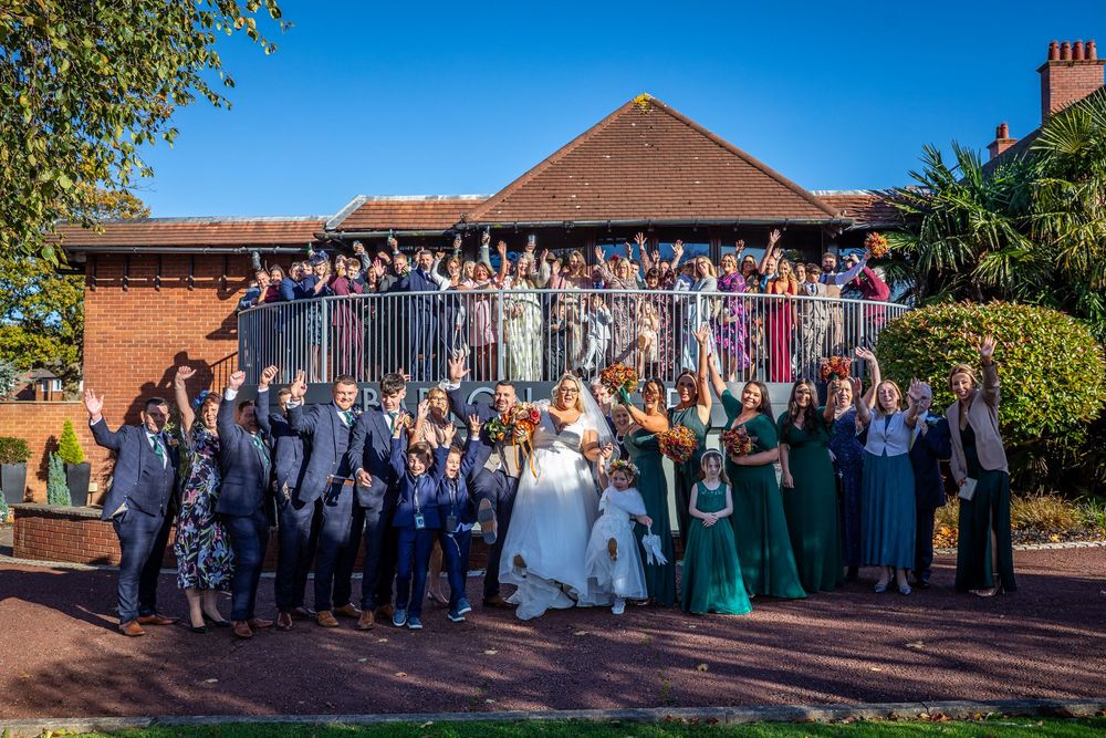 Large wedding party group photo with guests celebrating on balcony and grounds of brick venue.
