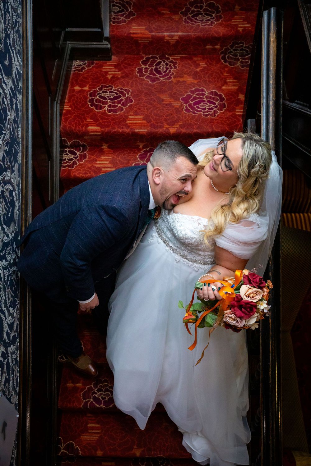 Bride and groom share a romantic kiss on red carpeted stairs, bride holding colorful autumn bouquet.
