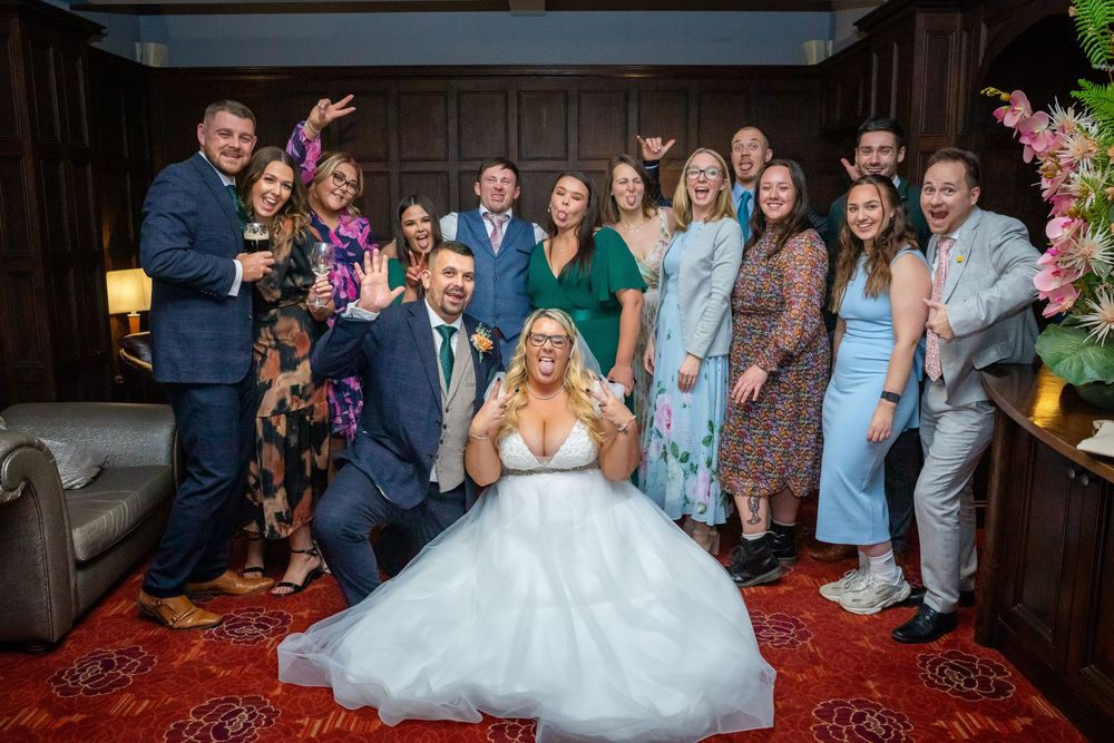 Bride in white gown poses with smiling wedding guests in elegant wood-paneled room with red carpet.