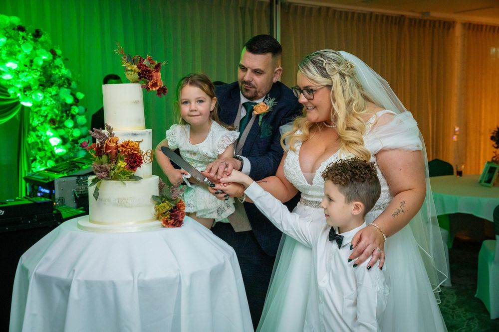Bride and groom cut wedding cake with two children beside them at elegant reception venue.
