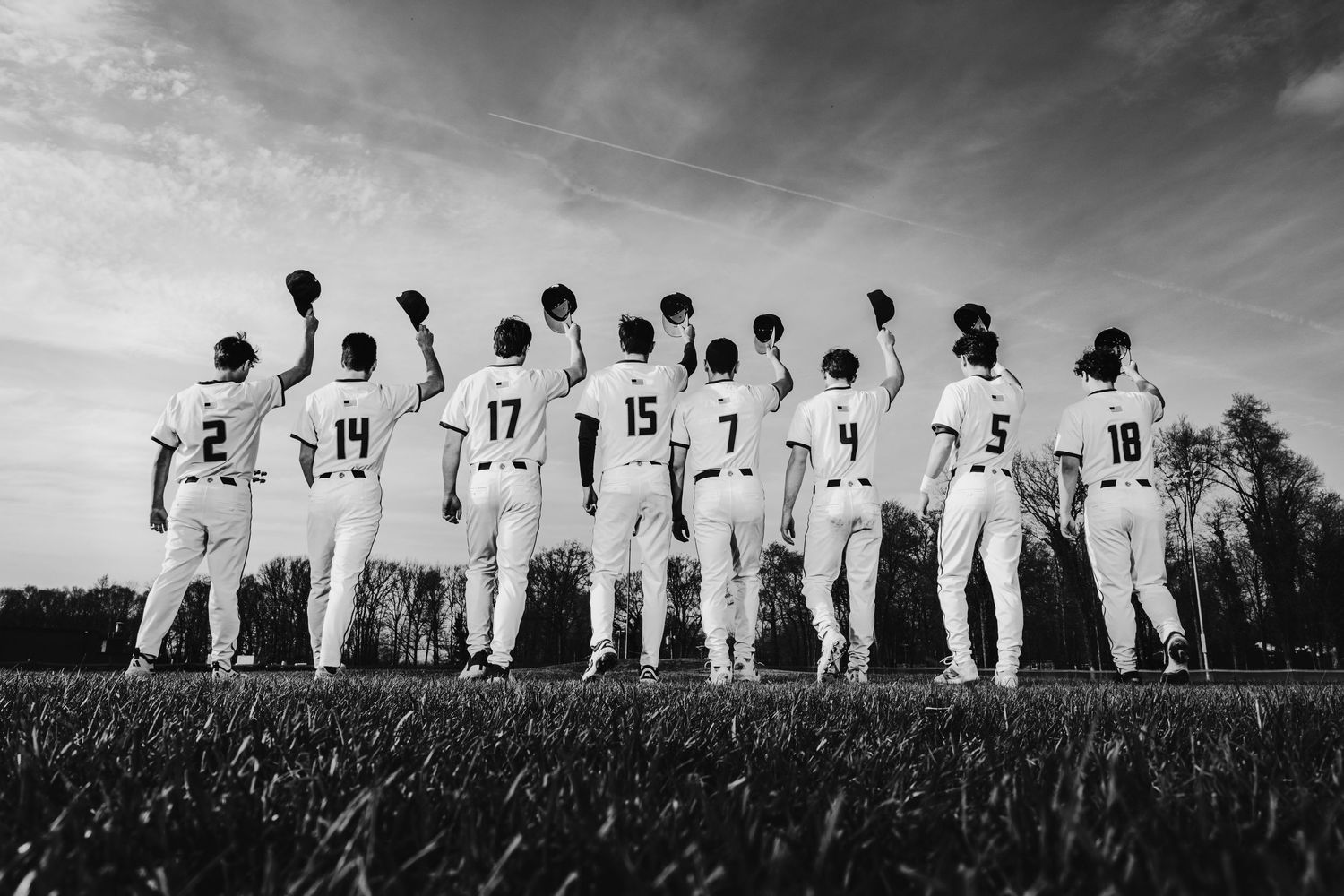 Black and white photo of a baseball team standing in a row from behind, raising their caps to the sky on a field.