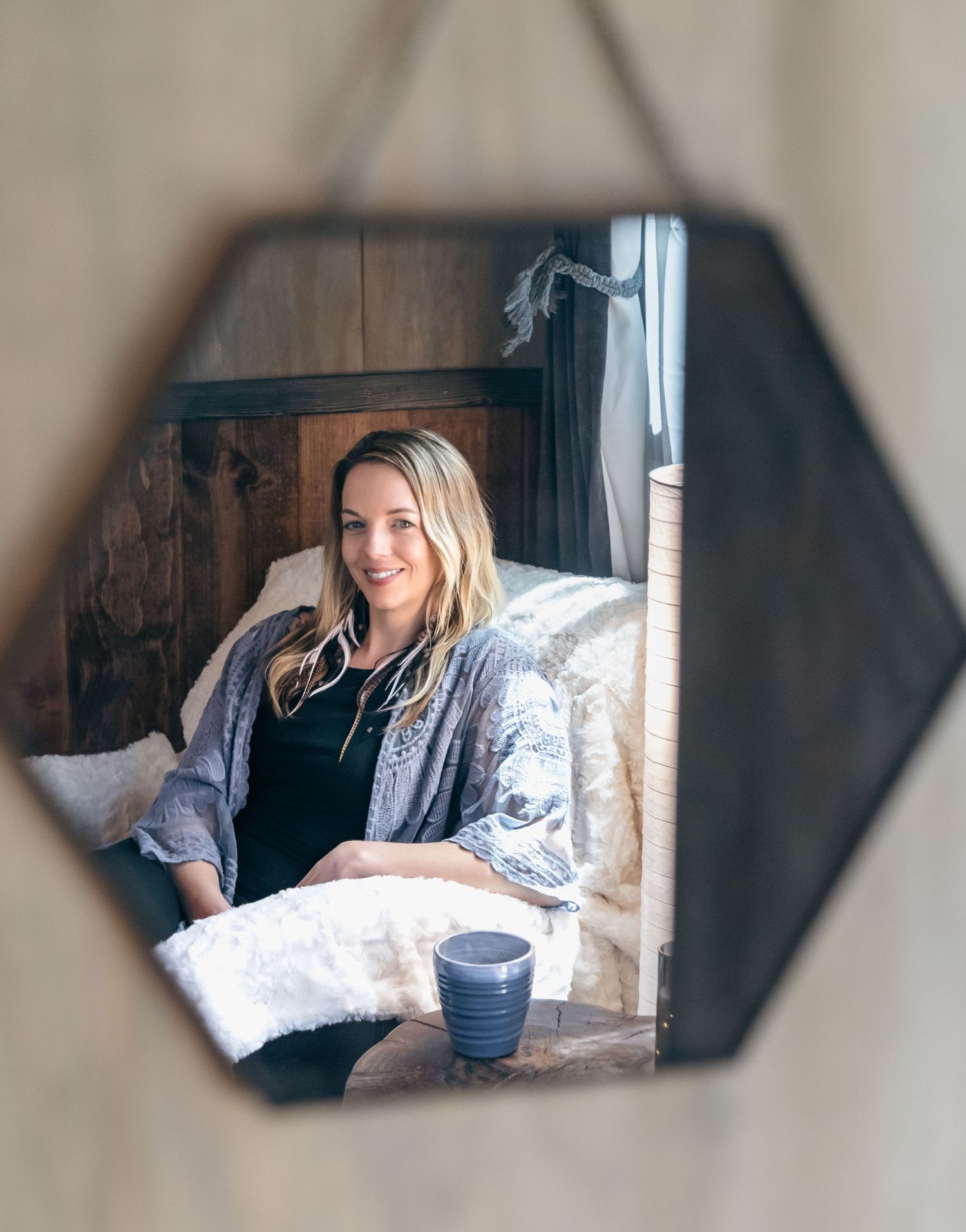 Woman smiling in a cozy rustic bedroom, reflected in a hexagonal hanging mirror with a mug nearby.