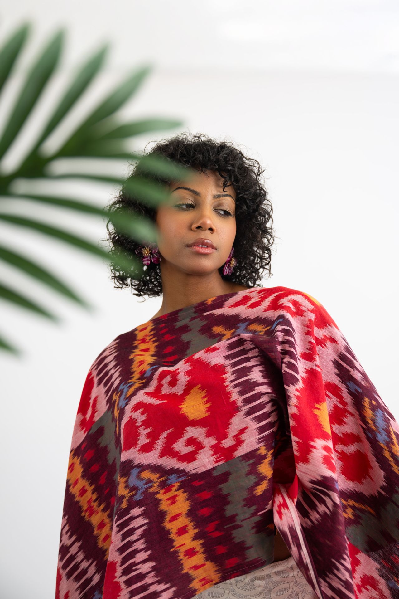 Woman in vibrant red ikat patterned wrap poses confidently before white background with tropical palm leaf.