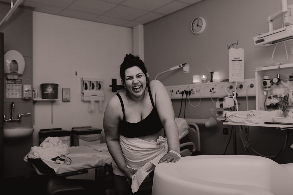 Black and white photo of a laughing woman in a hospital gown sitting in a medical room during labor.