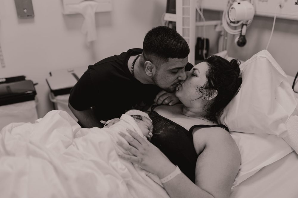 Black and white hospital photo of new parents sharing a tender kiss while mother holds their newborn baby in a delivery room.