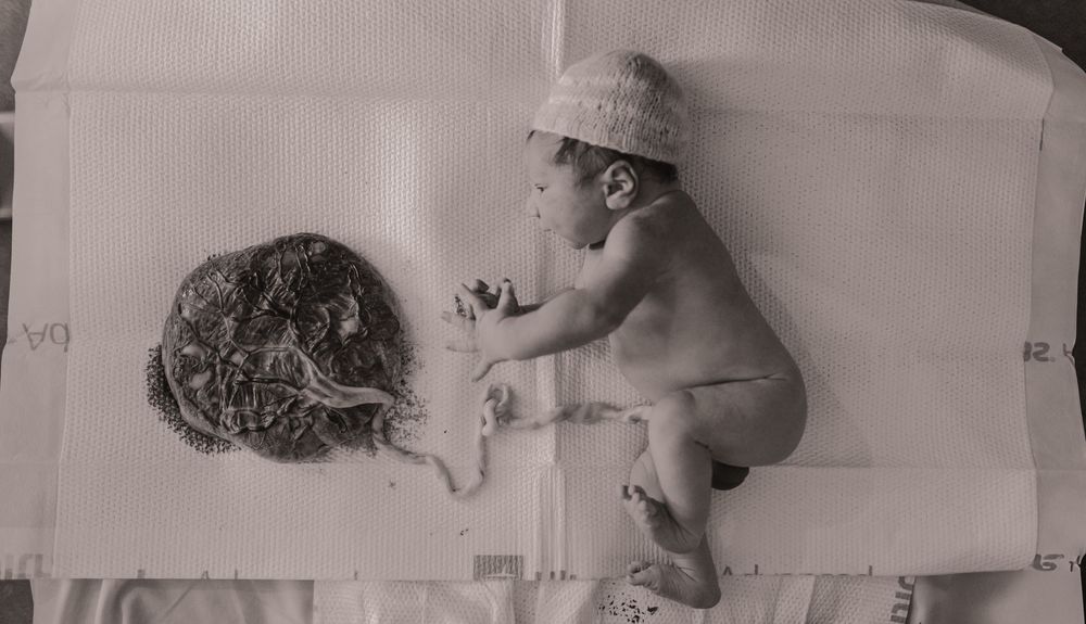 Newborn baby lying next to placenta on white surface in black and white birth photography.