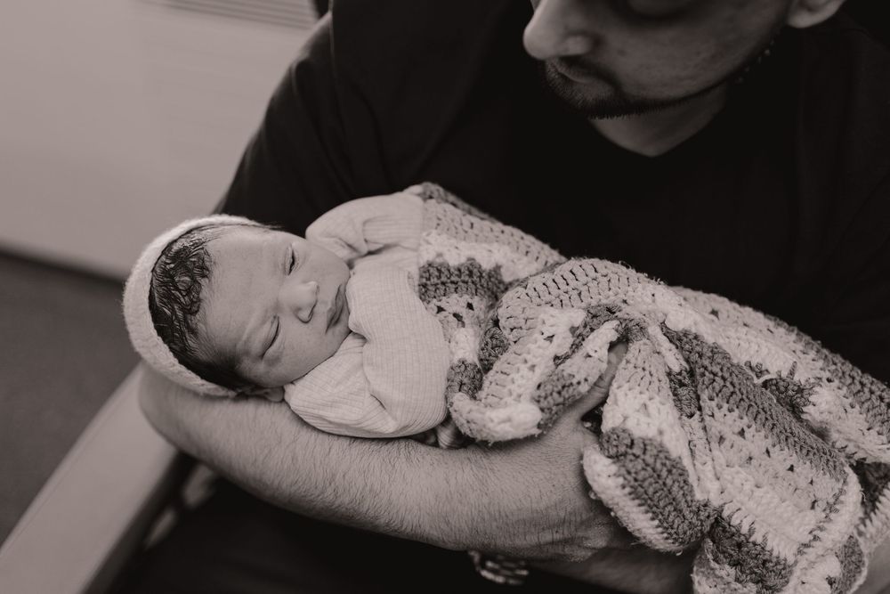 Black and white photo of a newborn baby sleeping peacefully, wrapped in a crochet blanket, cradled in a man's arms.