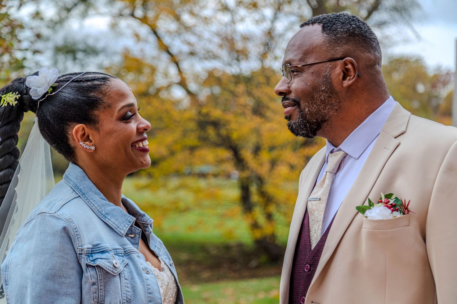 A bride in a white dress and denim jacket stands with a groom in a tan suit and burgundy vest outdoors.