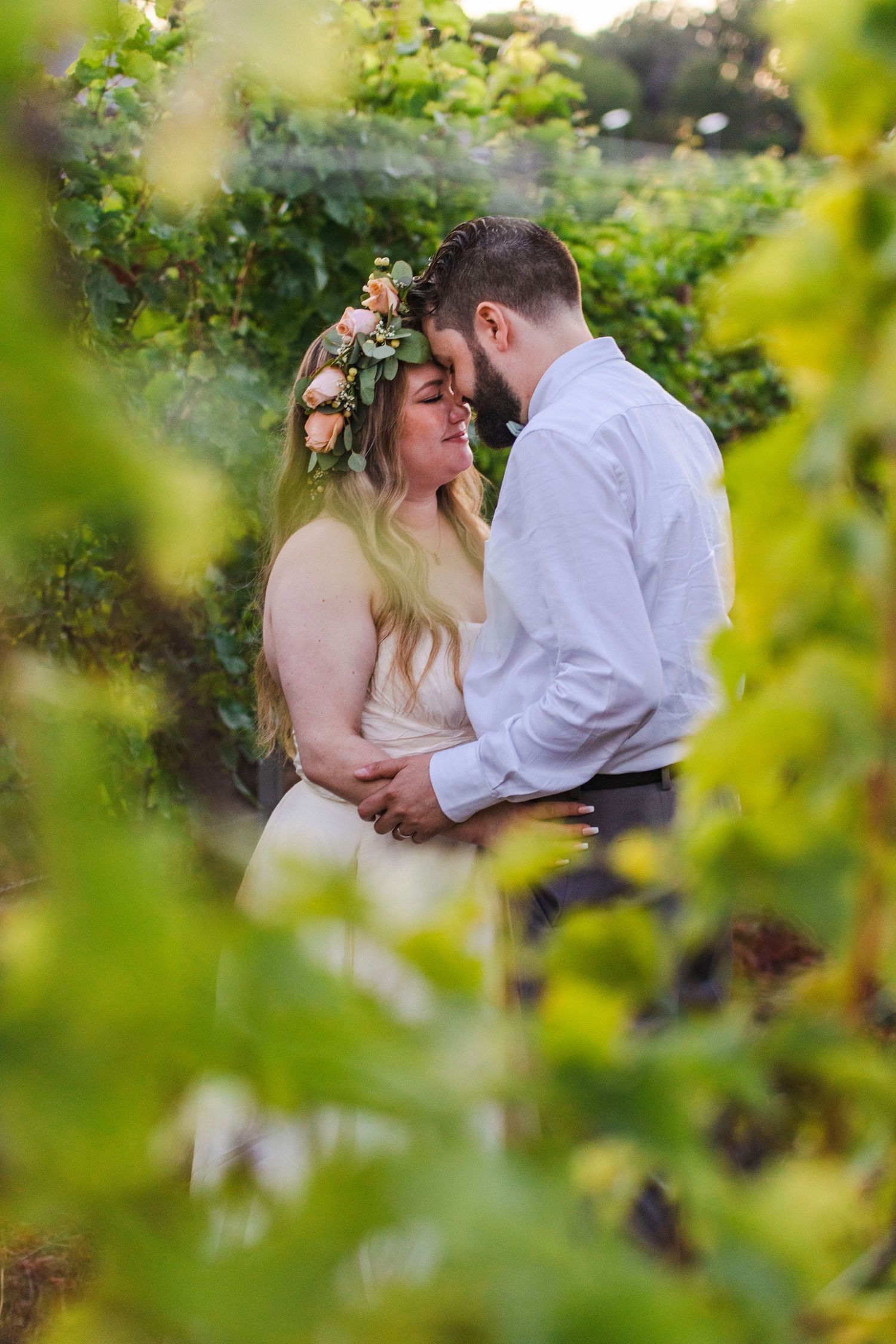 A couple shares intimate moments in a lush green vineyard setting wearing wedding attire and floral crown.