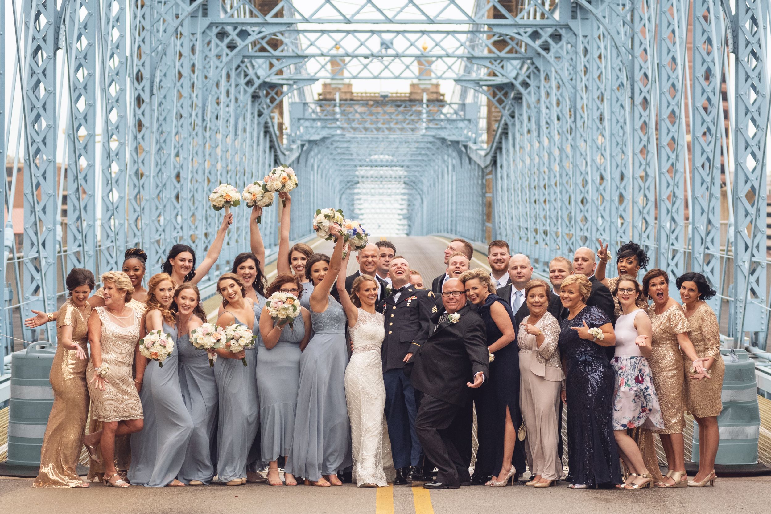 Large wedding party poses together on a blue bridge wearing coordinated formal attire in neutral tones.