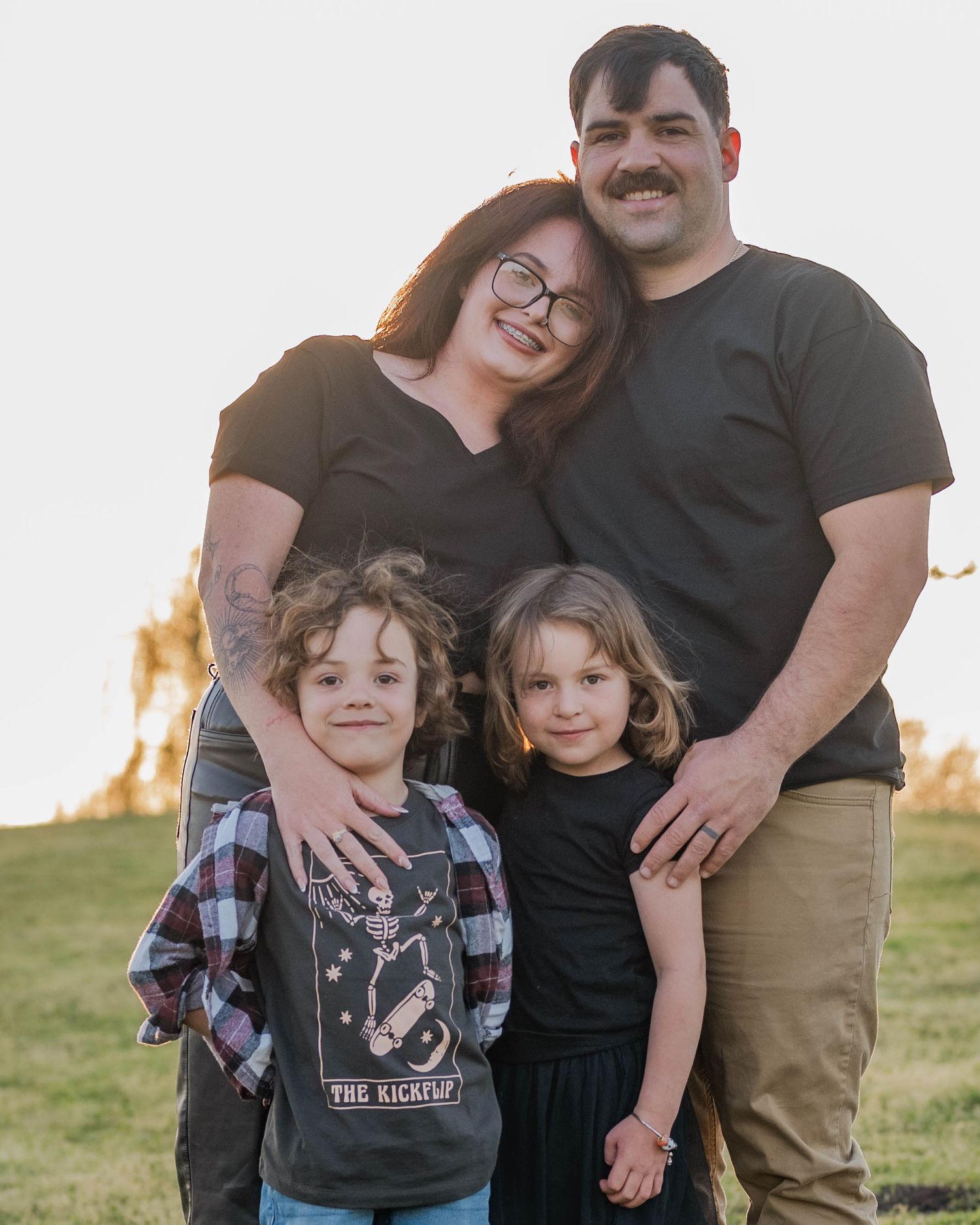A family of four poses together outdoors on a grassy hill during golden hour.