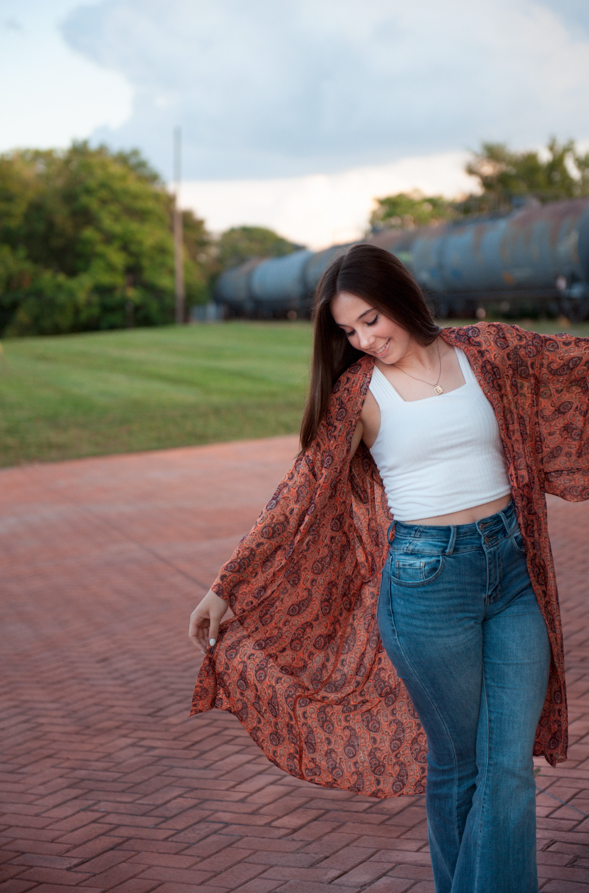 A person in jeans and white top poses playfully with a flowing floral kimono on a brick pathway outdoors.