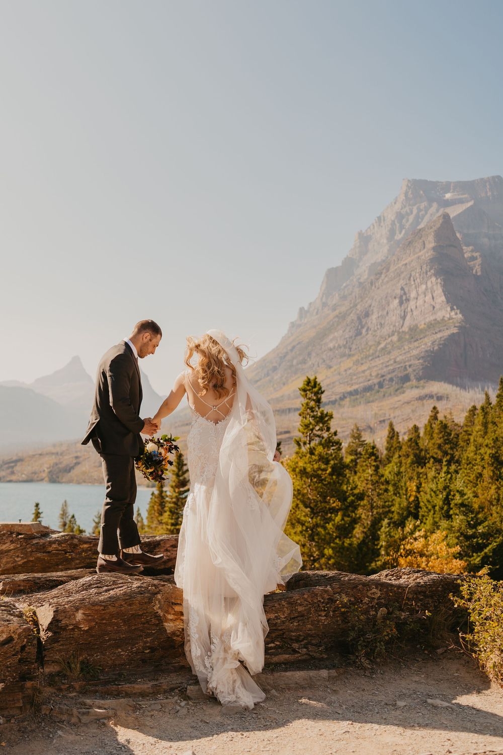 Glacier National Park Eloping Groom helps bride up onto rock on the windy day, captured by local Elopement Photographer Sabrina Marie