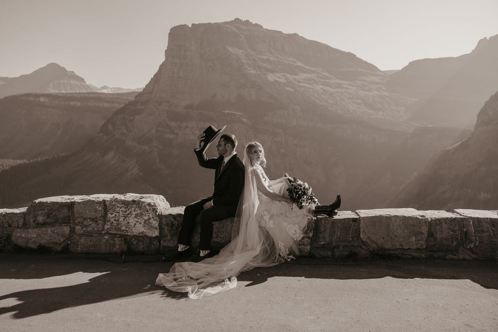 Glacier National Park Eloping Bride and Groom sitting along going to sun road putting on hat and looking at the surrounding mountains