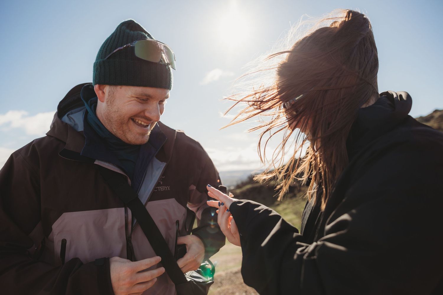 Two hikers sharing a phone outdoors on a sunny, windy day wearing winter jackets and beanies.