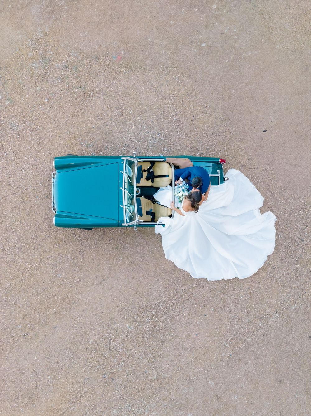 Aerial drone view of bride and groom riding in vintage teal convertible with flowing white gown.