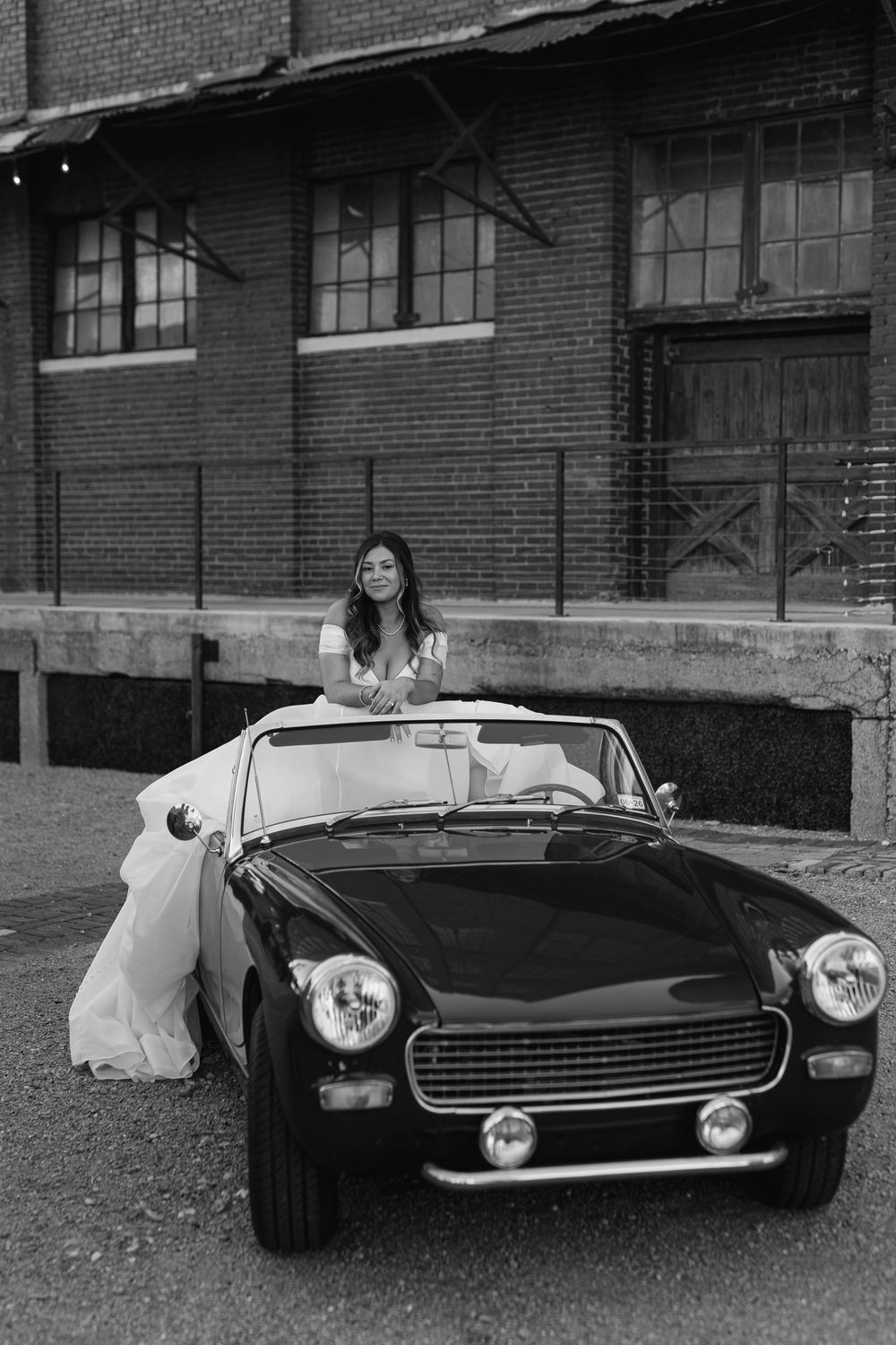 Black and white photo of a smiling bride in a white gown posing with a vintage convertible sports car.