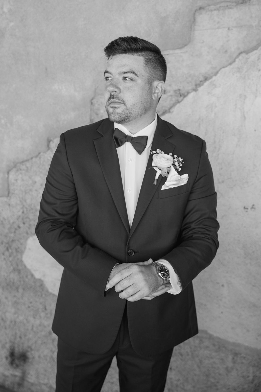 Dapper groom in black tuxedo with bow tie and boutonniere poses against textured wall.