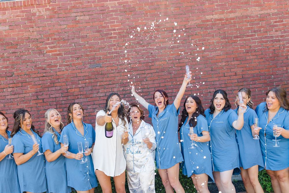 Bridesmaids in blue robes celebrate with bride popping champagne bottle against brick wall.