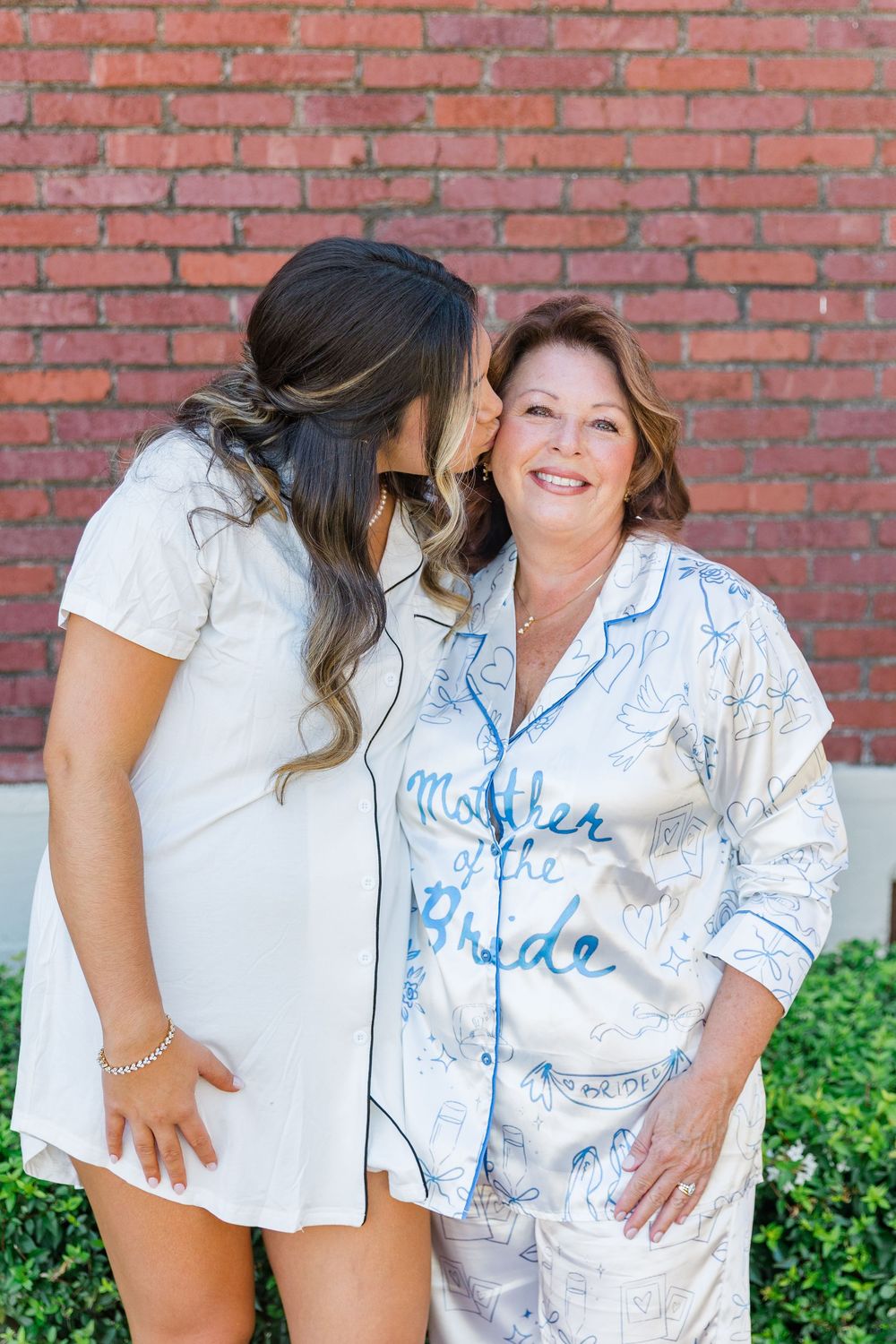 Bride kisses mother of the bride wearing matching satin pajamas in front of brick wall.
