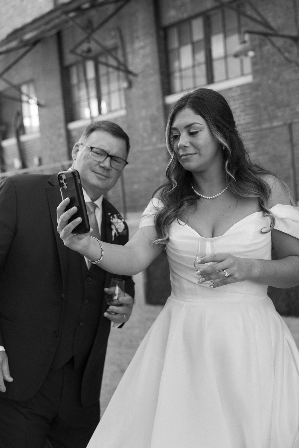 Bride in white gown and groom in suit take a selfie outdoors at wedding venue.