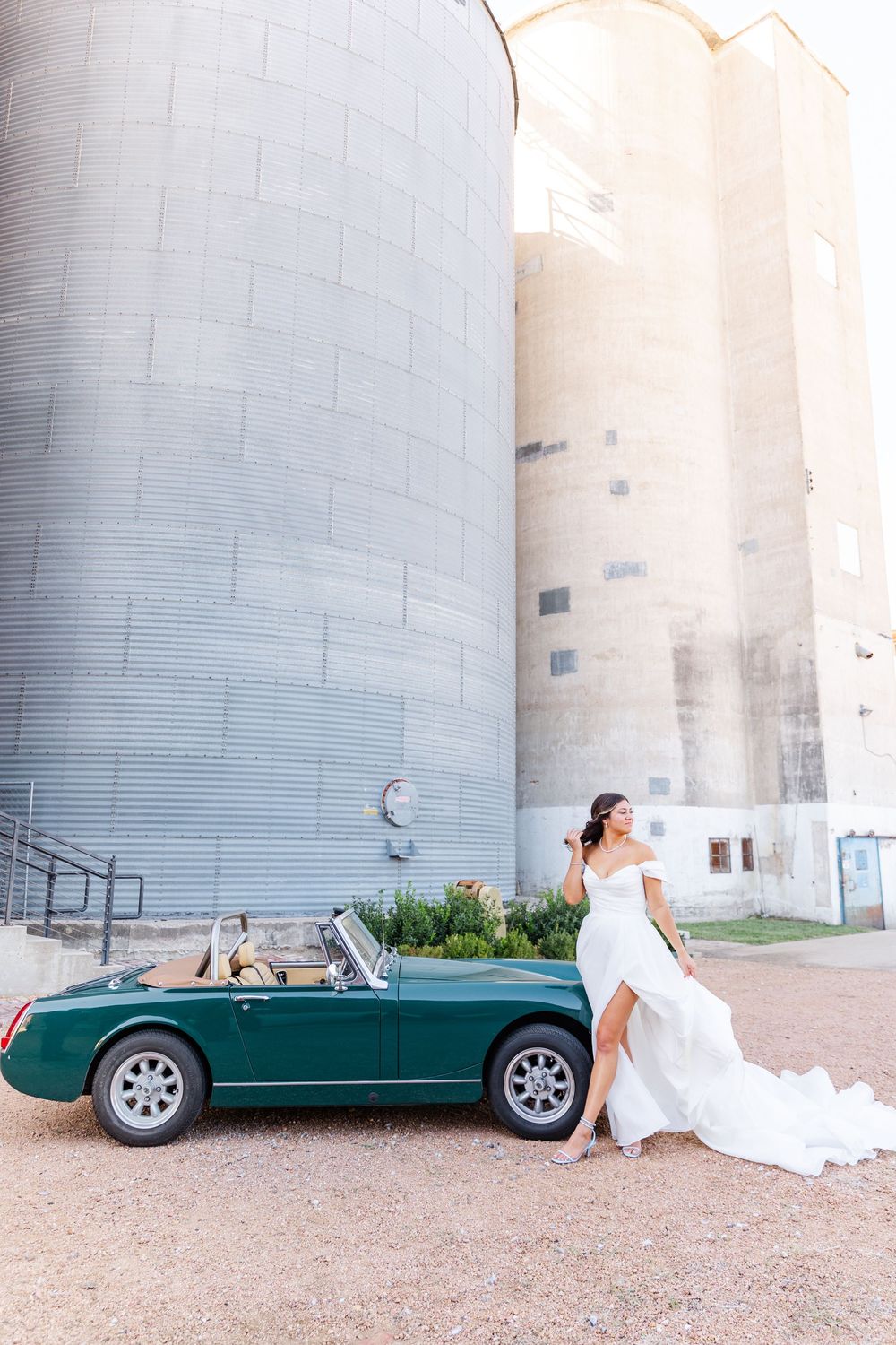 Bride in white off-shoulder gown leans against a green vintage convertible car beside industrial grain silos.
