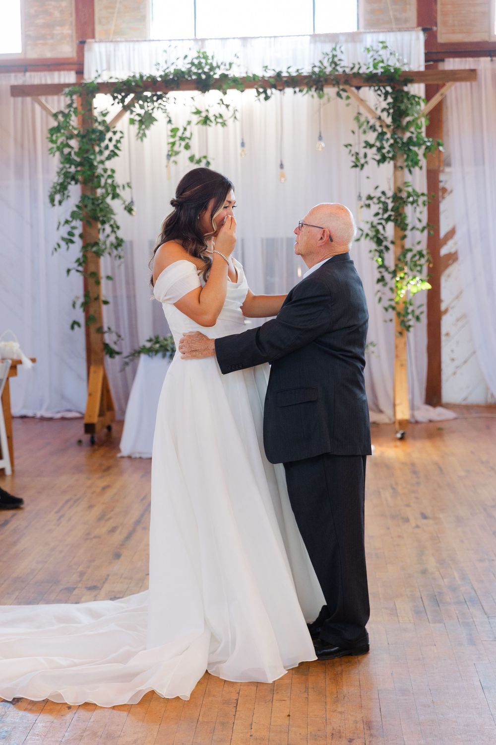 Emotional bride and father share first dance inside rustic wedding venue with floral arch.