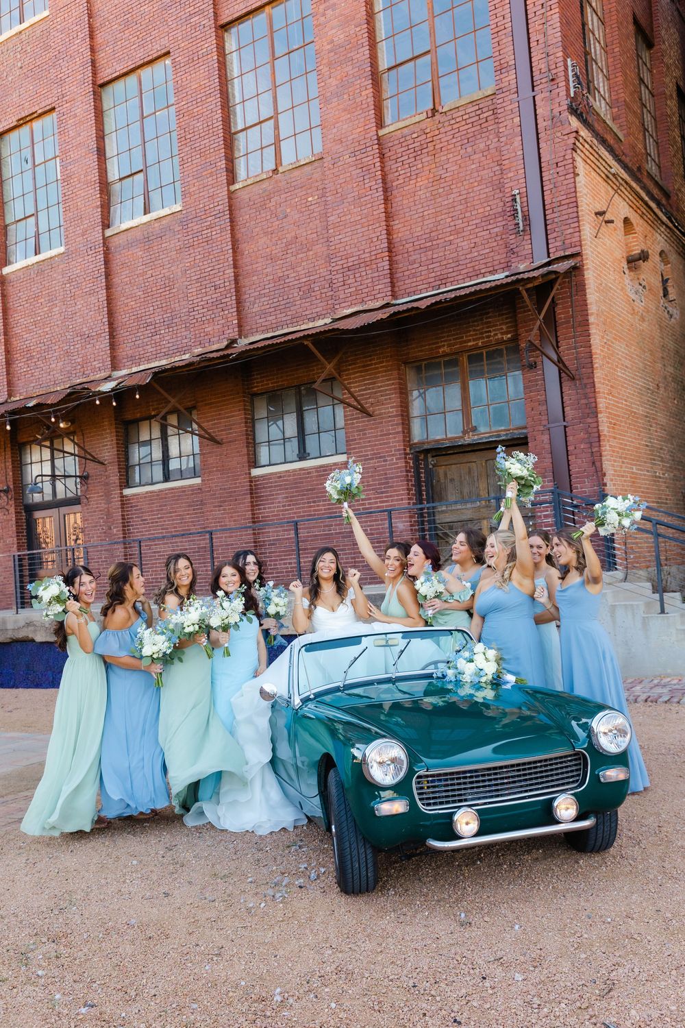 Bride and bridesmaids in blue gowns celebrate beside vintage teal convertible at brick building.